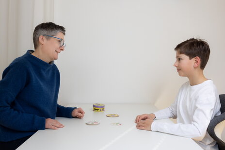 Ruth Vogel und ein Kind sitzen sich am Tisch gegenüber, spielen Dobble, sehen sich an und lächeln