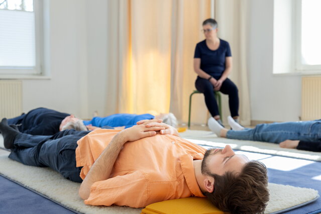 Eine Feldenkrais Gruppenstunde: Vier Menschen liegen am Boden. Ruth Vogel sitzt auf einem Hocker und leitet an. Die Menschen wirken entspannt